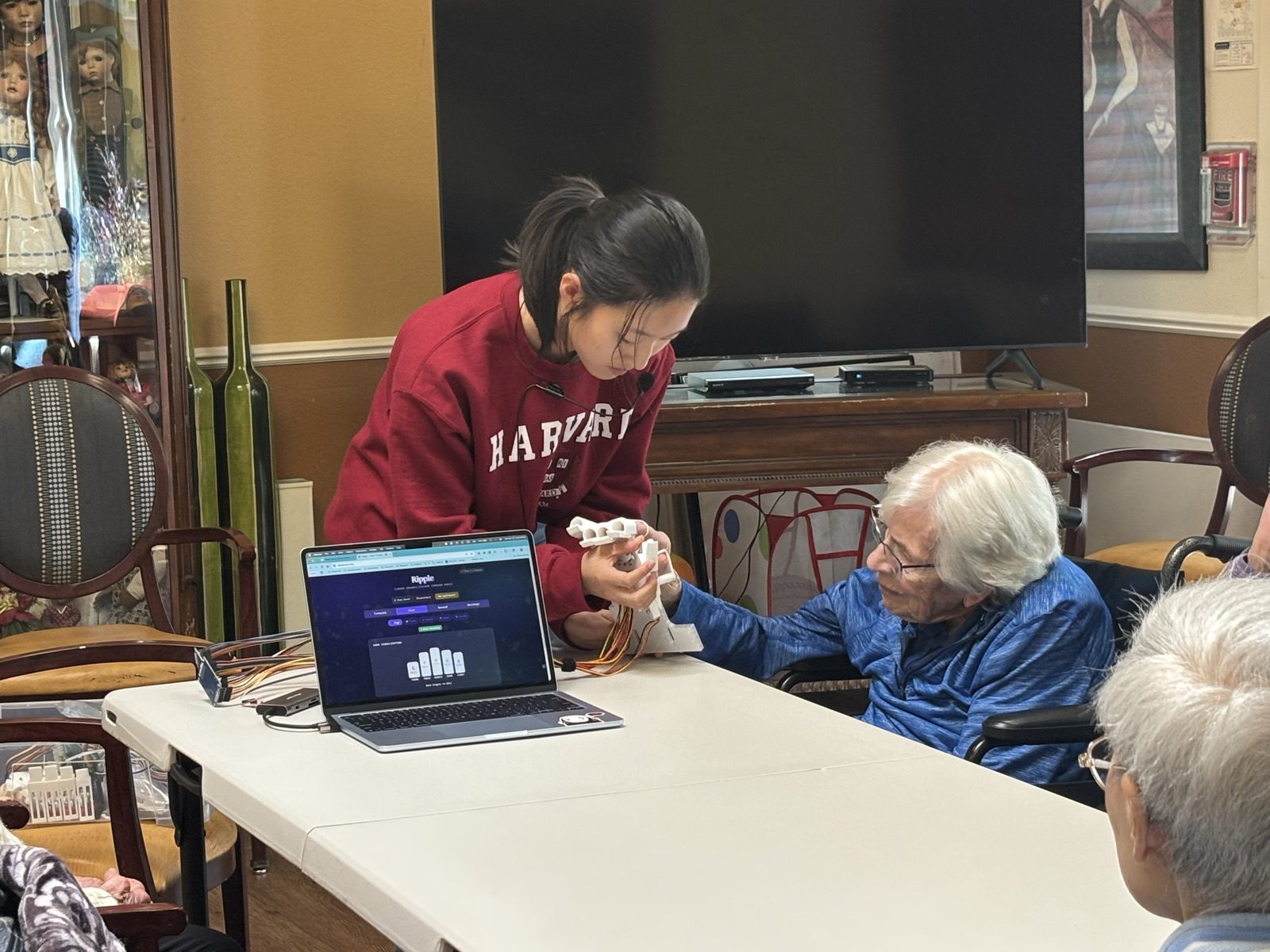 A resident wearing the Ripple device plays a note while Yanzi steadies their hand; the web app shows the live note bars on a laptop beside them